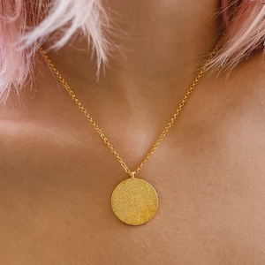 May include: A woman with pink hair wearing a white top with a delicate lace trim. She is wearing a gold necklace with a round pendant.