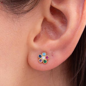 May include: A gold-colored flower-shaped stud earring with colorful gemstones. The earring features a central white gemstone surrounded by petals of blue, purple, red, green, and yellow gemstones. The earring is shown on an ear.