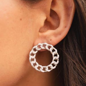 May include: Silver-toned chain link earrings. The earrings are circular, with a continuous chain design. The earrings are worn on the ear, and the image shows a close-up of the ear and the jewelry. The earrings are a simple yet stylish accessory.