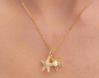 May include: A gold-colored necklace featuring two starfish pendants, one adorned with small clear crystals and the other with a small white pearl. The necklace is displayed against a cream-colored top.