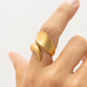 Op de afbeelding: Een goudkleurige ring met een open bypass-ontwerp, bestaande uit twee afzonderlijke, gladde en gebogen elementen die om de vinger wikkelen. Het metaal heeft een matte, geborstelde afwerking, wat zorgt voor een zachte, niet-reflecterende textuur op de band.