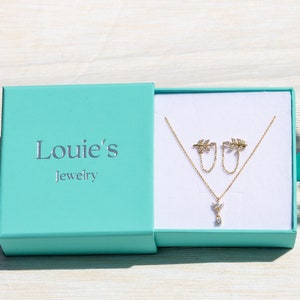 Pu&ograve; includere: Una scatola per gioielli turchese con il testo "Louie's Jewelry" sul coperchio. La scatola &egrave; aperta e contiene una collana d'oro con un piccolo ciondolo a forma di foglia e orecchini a foglia abbinati.