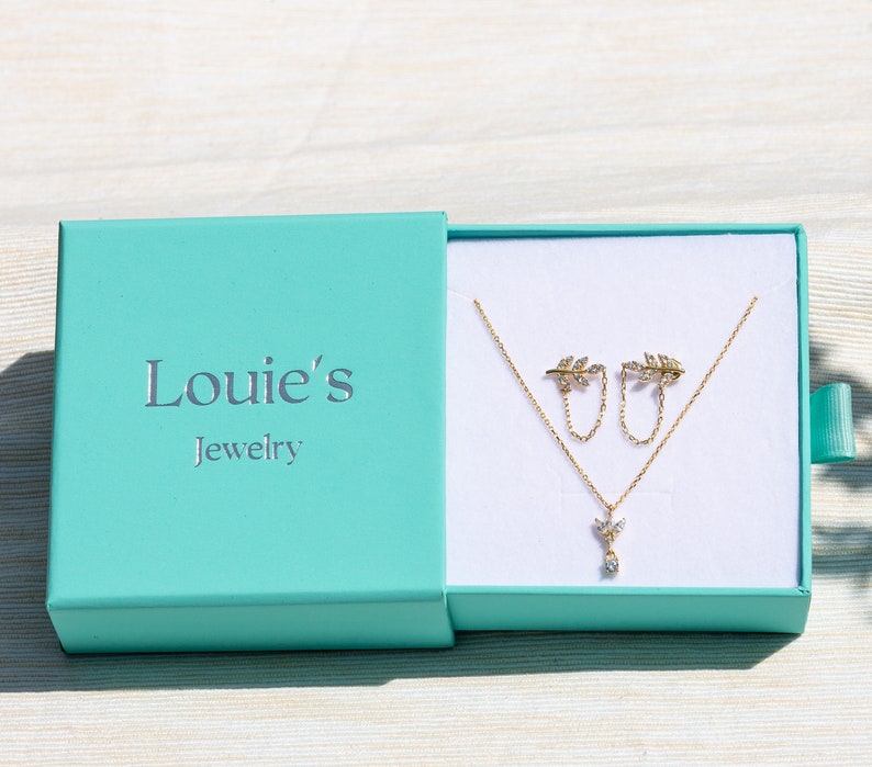 Pu&ograve; includere: Una scatola per gioielli turchese con il testo "Louie's Jewelry" sul coperchio. La scatola &egrave; aperta e contiene una collana d'oro con un piccolo ciondolo a forma di foglia e orecchini a foglia abbinati.