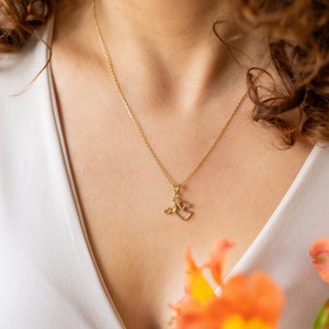 May include: A gold chain necklace with a small pendant featuring two intertwined hearts. The necklace is worn by a person with curly brown hair and a white top.