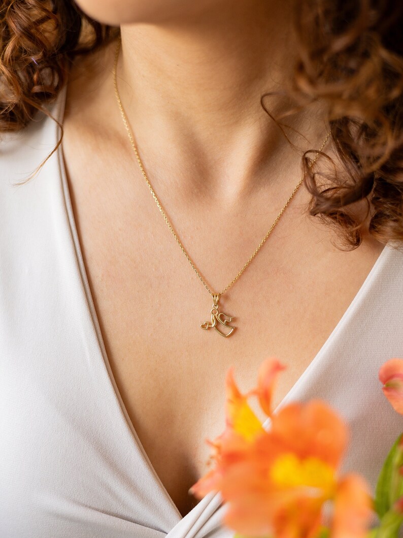 May include: A gold chain necklace with a small pendant featuring two intertwined hearts. The necklace is worn by a person with curly brown hair and a white top.