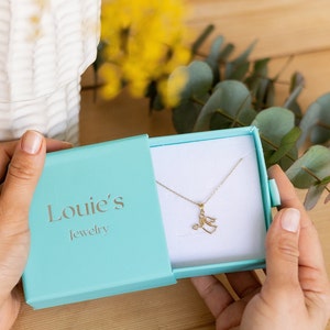 May include: A turquoise jewelry box with the text "Louie's Jewelry" on the lid. The box is open and a gold angel pendant necklace is inside.