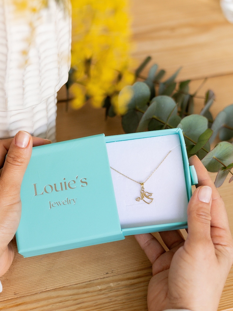 May include: A turquoise jewelry box with the text "Louie's Jewelry" on the lid. The box is open and a gold angel pendant necklace is inside.