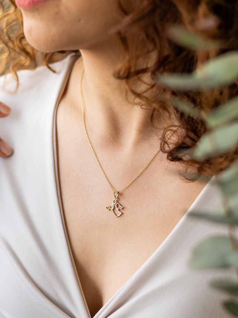 May include: A gold chain necklace with a delicate angel pendant. The angel is holding a small heart in its hand.