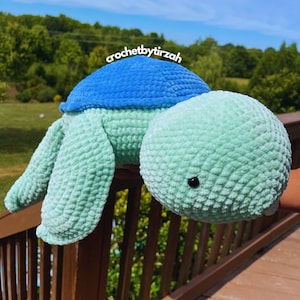 May include: A crocheted green turtle with a blue shell. The turtle is being held by a hand and is facing the camera. The background is a blurry image of a wooden deck and green trees.
