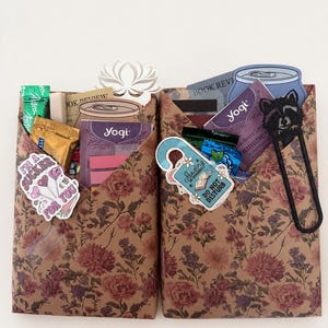 Puede incluir: Dos sobres de papel marrón con estampado floral que contienen varios artículos, incluyendo bolsitas de té Yogi, una pegatina que dice I'M A SUCKER FOR YOU, una pegatina de colgador de puerta READING IN PROGRESS DO NOT DISTURB y un clip de mapache negro.