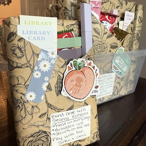 May include: A clear plastic bin filled with wrapped books and small gifts. The wrapping paper has a floral design. There are stickers, bookmarks, and library cards visible. One package has a handwritten note with the words "Blind Date w/a Steamy Romance."