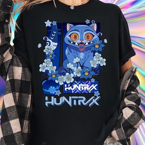 May include: Black t-shirt featuring a blue cartoon cat with large orange eyes, a pink tongue, and surrounded by blue flowers. The text "HUNTRAX" is displayed in white and blue. The shirt is worn by a person.