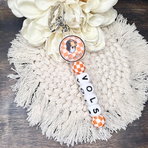 Peut inclure: Un porte-clés avec un charm rond représentant une image de dessin animé et le mot "Tennessee". Le porte-clés comprend des perles à carreaux orange et blanches épelant "VOLS". Le design incorpore des couleurs orange et blanc, avec un porte-clés argenté.