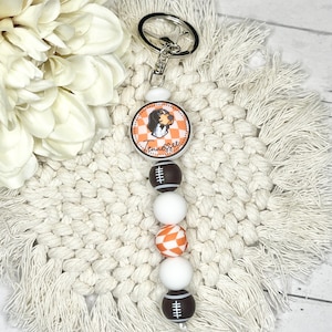 Puede incluir: Un amuleto a cuadros blanco y naranja con un perro y la palabra "Tennessee" en él, unido a un llavero plateado con una cadena de cuentas blancas, de fútbol marrón y cuentas blancas.