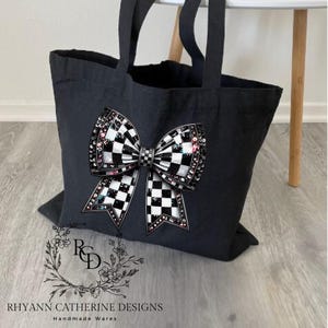 May include: Black canvas tote bag with a large checkered bow design. The bow is embellished with rhinestones and has a pink and white iridescent sheen. The bag has black straps and the text "RHYANN CATHERINE DESIGNS Handmade Wares" is printed on the bottom.