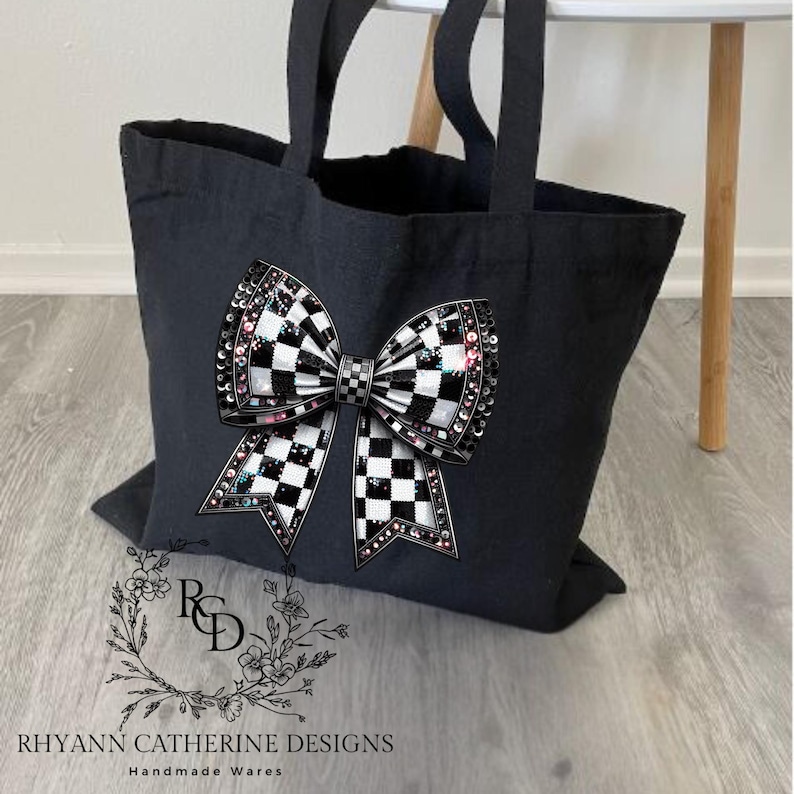 Puede incluir: Bolsa de lona negra con un gran dise&ntilde;o de lazo a cuadros. El lazo est&aacute; adornado con pedrer&iacute;a y tiene un brillo iridiscente rosa y blanco. La bolsa tiene asas negras y el texto "RHYANN CATHERINE DESIGNS Handmade Wares" est&aacute; impreso en la parte inferior.