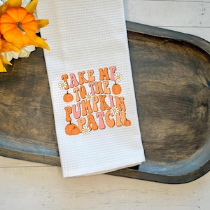 May include: White waffle-weave kitchen towel with the text "TAKE ME TO THE PUMPKIN PATCH" in orange, pink, and peach. The design includes pumpkins and daisies. The towel is displayed on a dark wooden tray.