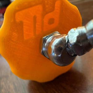 K&ouml;nnte beinhalten: Nahaufnahme eines orangefarbenen, strukturierten Knopfes mit den Buchstaben "TD" gepr&auml;gt. Der Knopf ist an einem silberfarbenen Metallanschluss befestigt. Das Bild ist leicht schr&auml;g aufgenommen und zeigt die Details des Knopfes und des Anschlusses.