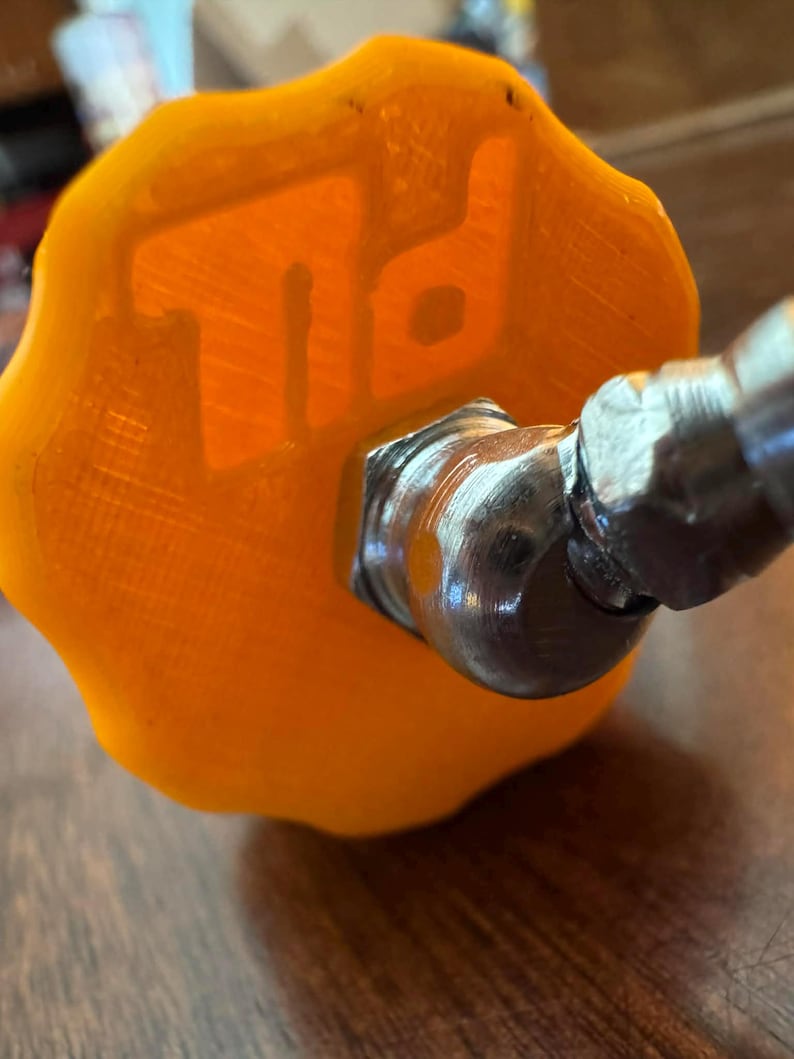 K&ouml;nnte beinhalten: Nahaufnahme eines orangefarbenen, strukturierten Knopfes mit den Buchstaben "TD" gepr&auml;gt. Der Knopf ist an einem silberfarbenen Metallanschluss befestigt. Das Bild ist leicht schr&auml;g aufgenommen und zeigt die Details des Knopfes und des Anschlusses.