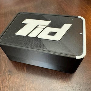 K&ouml;nnte beinhalten: Eine schwarze rechteckige Box mit wei&szlig;em Deckel. Der Deckel tr&auml;gt die wei&szlig;en Buchstaben "Tid". Die Box steht auf einer Holzoberfl&auml;che.