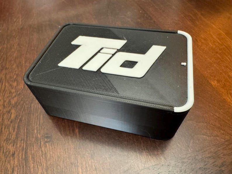 K&ouml;nnte beinhalten: Eine schwarze rechteckige Box mit wei&szlig;em Deckel. Der Deckel tr&auml;gt die wei&szlig;en Buchstaben "Tid". Die Box steht auf einer Holzoberfl&auml;che.