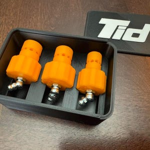 K&ouml;nnte beinhalten: Drei orangefarbene, zylindrische Objekte mit silbernen Armaturen sind in einem schwarzen rechteckigen Beh&auml;lter angeordnet. Ein schwarzes rechteckiges Objekt mit den wei&szlig;en Buchstaben "Tid" befindet sich neben dem Beh&auml;lter. Die Objekte befinden sich auf einer Holzoberfl&auml;che.