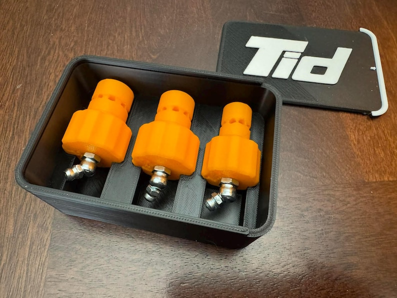 K&ouml;nnte beinhalten: Drei orangefarbene, zylindrische Objekte mit silbernen Armaturen sind in einem schwarzen rechteckigen Beh&auml;lter angeordnet. Ein schwarzes rechteckiges Objekt mit den wei&szlig;en Buchstaben "Tid" befindet sich neben dem Beh&auml;lter. Die Objekte befinden sich auf einer Holzoberfl&auml;che.