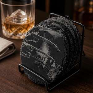 Könnte beinhalten: Ein Set schwarzer Schieferuntersetzer mit strukturierter Oberfläche in einem schwarzen Metallständer. Die runden Untersetzer scheinen aus Stein zu sein. Im Hintergrund ist ein Glas Whisky mit einem großen Eiswürfel zu sehen.