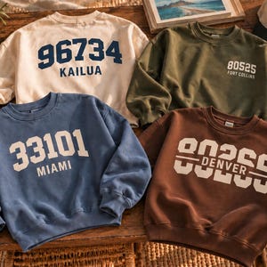 Pode incluir: Quatro camisolas de gola redonda em creme, verde azeitona, azul e castanho. A camisola creme apresenta o texto azul-marinho 96734 KAILUA. A verde azeitona tem o texto branco 80525 FORT COLLINS. A azul tem o texto branco 33101 MIAMI. A castanha tem 80206 DENVER.