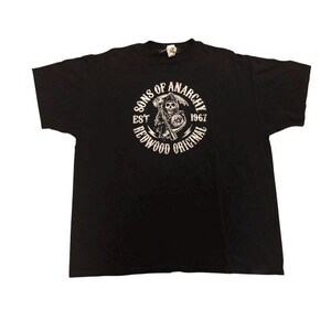 Peut inclure: Un t-shirt noir à manches courtes avec un motif circulaire blanc sur la poitrine. Le design représente une faucheuse tenant une faux et le texte SONS OF ANARCHY EST 1967 REDWOOD ORIGINAL disposé en cercle autour de la figure centrale.
