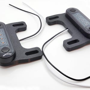 May include: Two black LED light assemblies with mounting brackets. Each assembly has a rectangular light housing with five visible LEDs and two black buttons. Black and white wires extend from each unit.