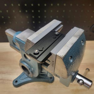 May include: A black metal sight is clamped in a blue metal vise. The vise is labeled "ARC4".