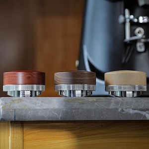 May include: Three wooden coffee machine portafilter handles with silver metal bases. The wood colors vary from dark to light, showcasing the natural grain. The handles sit on a gray marble surface.