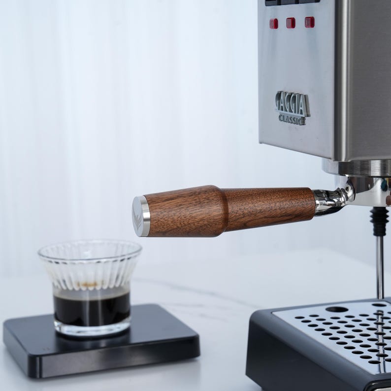 May include: A stainless steel espresso machine with a wooden handle and a small glass cup of coffee. The machine has the brand name "Gaggia Classic" on the front. The cup of coffee sits on a small black square.