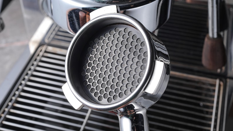 May include: A close-up of a silver espresso portafilter with a black, honeycomb-shaped filter basket.