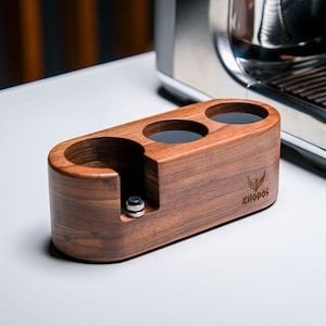 May include: A wooden espresso cup holder with three slots and a compartment for a tamper. The holder is made of walnut wood and has the word "Knodos" engraved on the side.