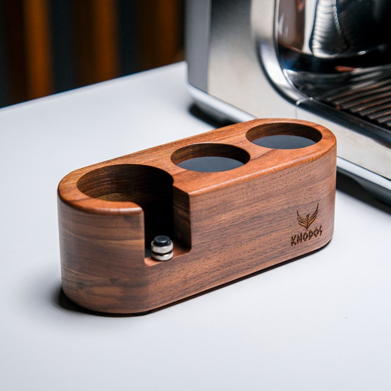 May include: A wooden espresso cup holder with three slots and a compartment for a tamper. The holder is made of walnut wood and has the word "Knodos" engraved on the side.