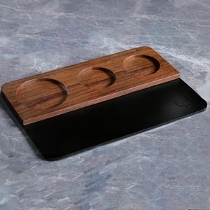 Puede incluir: Una bandeja de madera con tres hendiduras circulares para sostener tazas o vasos. La bandeja está hecha de madera oscura y tiene una base negra.