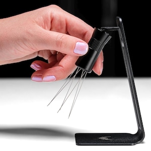 May include: A black metal stand with a small black tool with multiple thin metal needles extending from the bottom. The tool is being held by a hand.