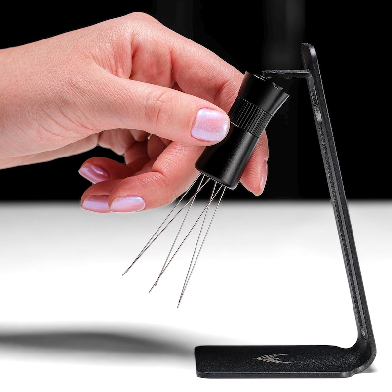 May include: A black metal stand with a small black tool with multiple thin metal needles extending from the bottom. The tool is being held by a hand.
