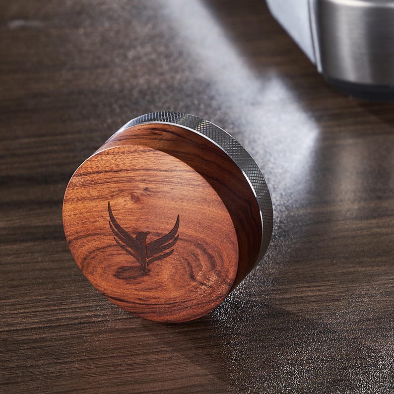May include: A round wooden tamper with a silver metal band. The tamper has a bird logo engraved on the top.