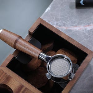 May include: A wooden espresso tamper holder with a stainless steel tamper and a black interior. The holder has a compartment for storing coffee grounds.