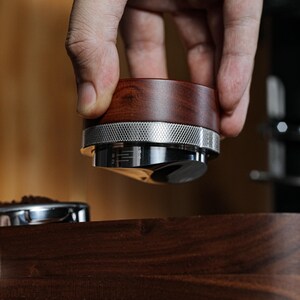 May include: A close-up of a wooden and metal coffee tamper being held over a coffee portafilter. The tamper has a wooden handle and a metal base with a ring around it. The portafilter is sitting on a wooden surface.