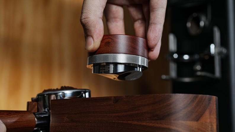 May include: A close-up of a wooden and metal coffee tamper being held over a coffee portafilter. The tamper has a wooden handle and a metal base with a ring around it. The portafilter is sitting on a wooden surface.