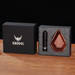 Puede incluir: Una caja negra con el logotipo "KNODOS" y un diseño de pájaro plateado. La caja contiene una botella negra y un cuenco de madera.