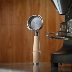 May include: A stainless steel portafilter with a light wood handle, for espresso machines. The portafilter is in front of a black espresso machine, with a wooden background.