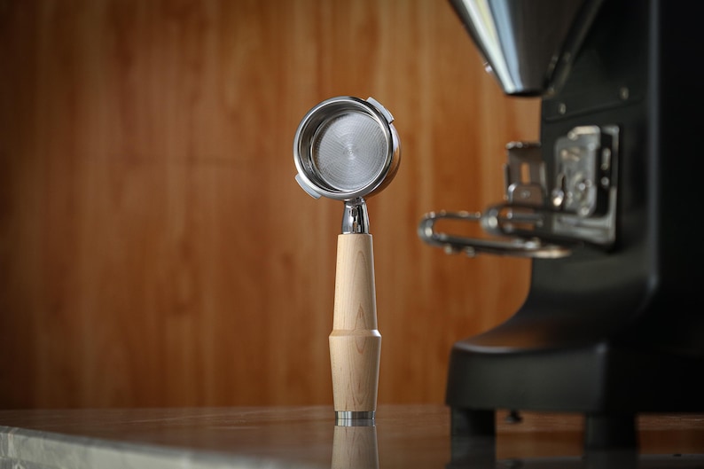 May include: A stainless steel portafilter with a light wood handle, for espresso machines. The portafilter is in front of a black espresso machine, with a wooden background.