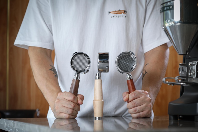 May include: Three espresso machine portafilters with wooden and metal handles are held in front of a person wearing a white t-shirt with a Patagonia logo. The portafilters are displayed against a blurred background of a coffee machine.