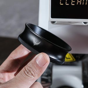 May include: A black metal portafilter with a silver logo, held in a hand.