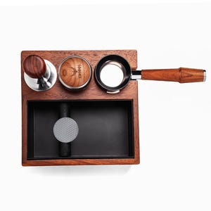 Puede incluir: Una estación de espresso de madera con un tamper, un distribuidor, un portafiltro y un cajón para golpear. La estación es de madera oscura y presenta un tamper y un portafiltro de acero inoxidable. El cajón para golpear tiene una almohadilla de goma gris.